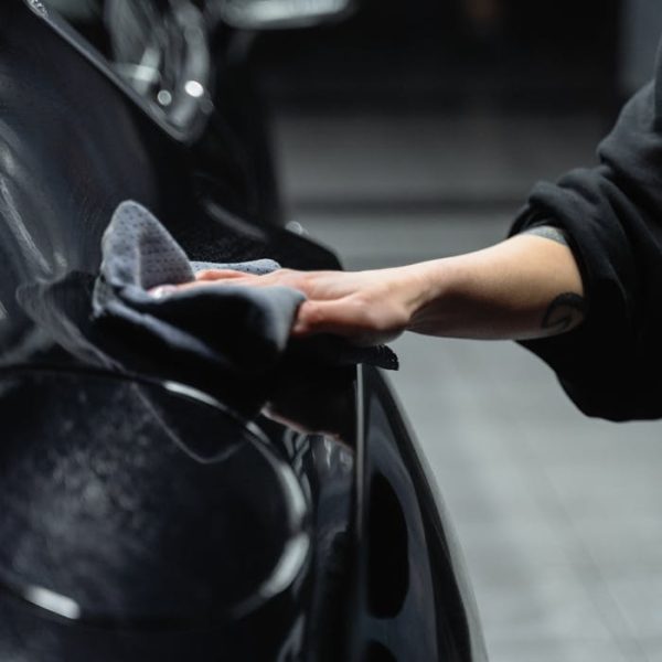 Close-up of a person's hand cleaning a shiny black car with a microfiber cloth in a garage.
