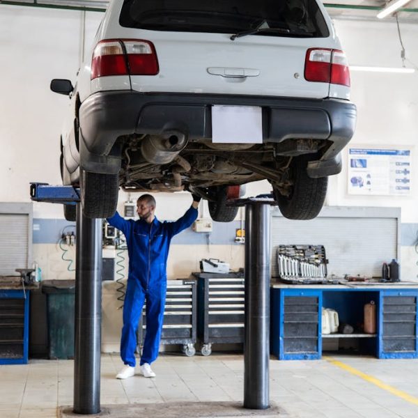 Mechanic in blue coverall inspecting a lifted car in an auto repair shop.