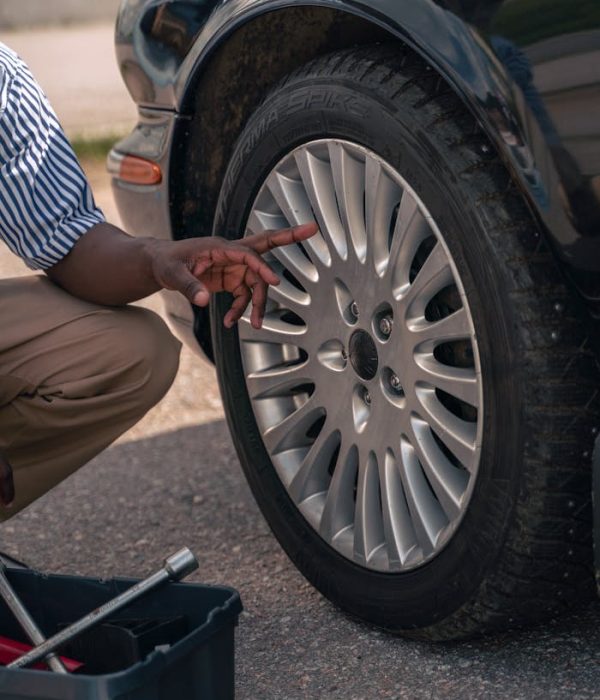 An adult male pointing at a car tire, suggesting a focus on vehicle maintenance or inspection outdoors.
